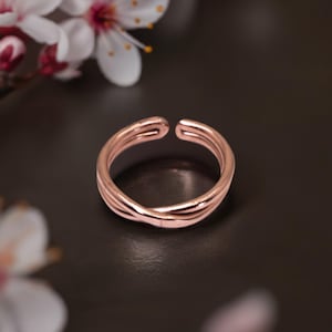 May include: A rose gold-toned open ring featuring a double-band design that crosses over at the front. The metal has a smooth, polished finish with a gap at the top, allowing for adjustable sizing. The ring is displayed against a dark background.