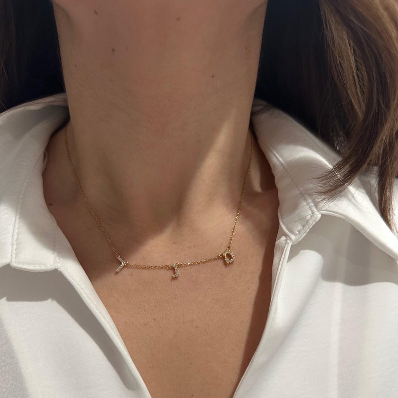 May include: A gold chain necklace with three small, diamond-encrusted letters hanging from it.