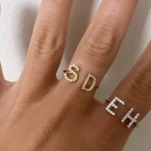 以下が含まれることがあります： クリアなキュービックジルコニアで飾られた「SD」と「EH」の文字が刻まれた2つの金のリング。リングは人の指につけられています。