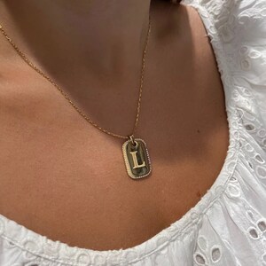 May include: A gold chain necklace with a gold pendant in the shape of a rectangular tag with a raised letter L in the center. The tag has a diamond-like border.