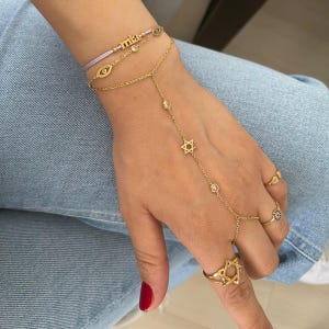 May include: A gold chain bracelet with a Star of David charm and a small eye charm. The bracelet is layered with a string bracelet with the word "mia" and a small eye charm.