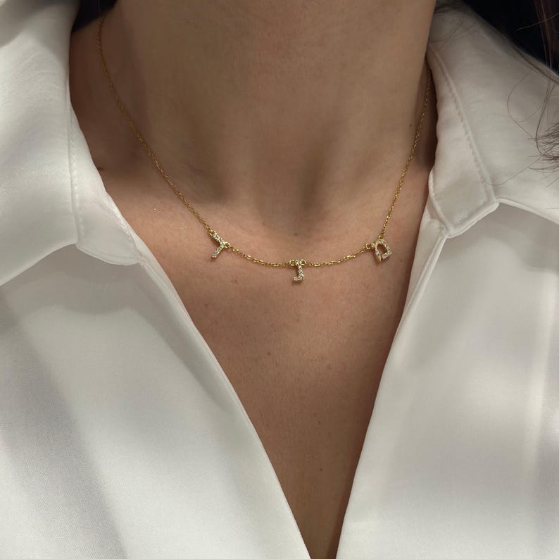 May include: A gold chain necklace with three small charms, each featuring a letter set with clear cubic zirconia stones.
