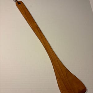 May include: A wooden spatula with a long handle and a wide, flat head. The spatula is made of light brown wood and has a leather loop for hanging. The spatula is approximately 12 inches long.