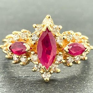 May include: A gold ring featuring a large central marquise-cut red ruby flanked by two smaller marquise-cut rubies. The setting is accented with numerous small round white diamonds arranged in a cluster around the three primary red gemstones.