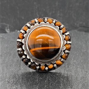 May include: A silver ring with a large round tiger's eye stone set in a silver bezel. The bezel is surrounded by a ring of smaller tiger's eye stones.