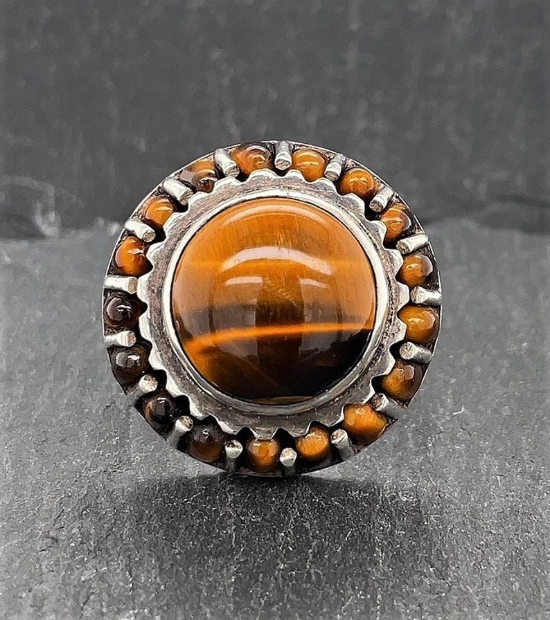 May include: A silver ring with a large round tiger's eye stone set in a silver bezel. The bezel is surrounded by a ring of smaller tiger's eye stones.