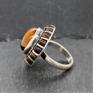 May include: A silver ring with a large oval tiger's eye stone set in a bezel setting. The ring is surrounded by a band of smaller tiger's eye stones.