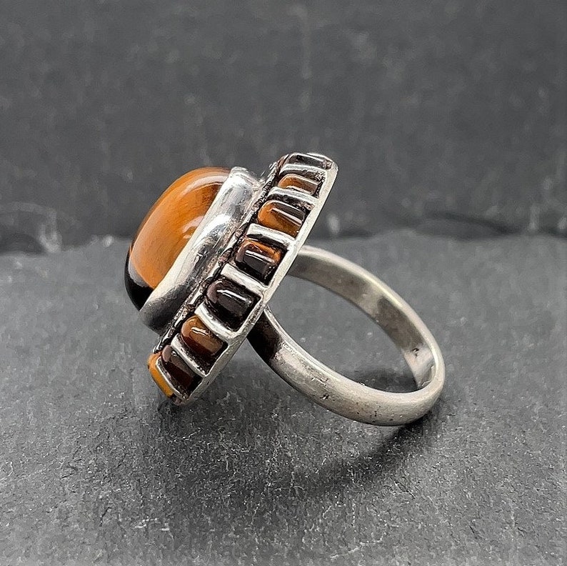 May include: A silver ring with a large oval tiger's eye stone set in a bezel setting. The ring is surrounded by a band of smaller tiger's eye stones.