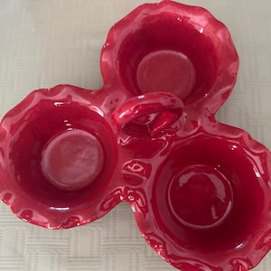 May include: A glossy red ceramic serving dish featuring three interconnected circular bowls with ruffled, wavy edges. A central arched handle connects the three bowls, providing a unified structure for this bright red kitchen serving piece.