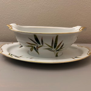 May include: A white porcelain gravy boat with an attached underplate, featuring a delicate bamboo leaf pattern in shades of green and brown. The rim of both the boat and the plate is accented with a thin, elegant gold trim along the edges.