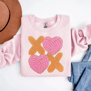 Könnte beinhalten: Ein hellrosa Sweatshirt mit einem Design aus zwei herzförmigen rosa Conchas und zwei orangefarbenen X'en. Die Conchas haben ein strukturiertes Muster, und die X'e bestehen aus Holzbrettern. Das Sweatshirt ist ein Rundhals-Stil.