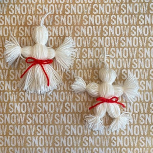 May include: Two white yarn snowmen ornaments with red yarn bows. The snowmen are on a brown background with the word "snow" repeated.