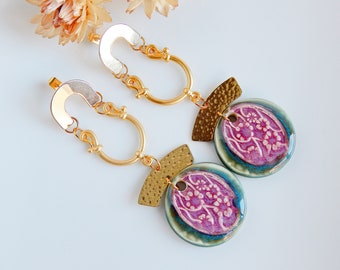May include: A pair of gold earrings with a pink and green ceramic pendant featuring a floral design. The earrings have a gold hoop and a gold chain with a small metal charm.