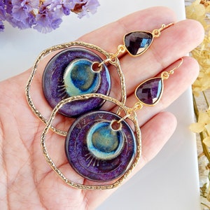 Op de afbeelding: Paarse en blauwe keramische oorbellen met gouden hoepels en druppelvormige amethiststenen.