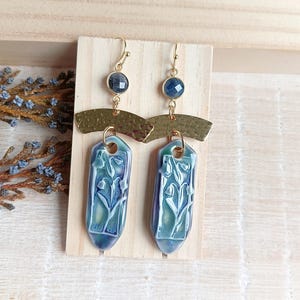 Puede incluir: Un par de pendientes de cerámica azul y verde con un diseño floral. Los pendientes están suspendidos de aros de metal dorado con detalles de piedras preciosas azules.