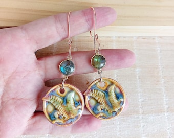 May include: A pair of handmade earrings with a unique design. Each earring features a round, ceramic pendant with a raised, textured pattern in shades of yellow, blue, and green. The earrings also have a small, round, iridescent stone and are suspended from a copper-colored hook.