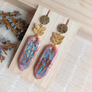 May include: A pair of dangle earrings with a floral design. The earrings are made of ceramic and have a purple, blue, and brown color scheme. The earrings are accented with gold-colored metal.