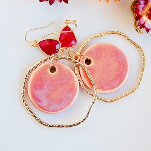 Peut inclure: Une paire de boucles d'oreilles cr&eacute;oles dor&eacute;es avec des cercles en c&eacute;ramique rose qui pendent. Les cr&eacute;oles ont une finition textur&eacute;e et les cercles en c&eacute;ramique ont un effet marbr&eacute; subtil. Les boucles d'oreilles sont rehauss&eacute;es d'accents en pierres pr&eacute;cieuses rouges.