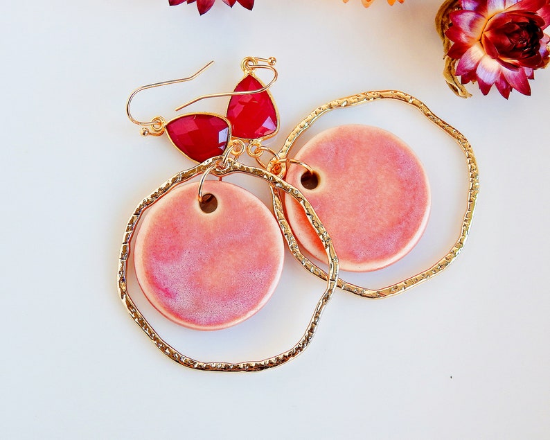 Peut inclure: Une paire de boucles d'oreilles cr&eacute;oles dor&eacute;es avec des cercles en c&eacute;ramique rose qui pendent. Les cr&eacute;oles ont une finition textur&eacute;e et les cercles en c&eacute;ramique ont un effet marbr&eacute; subtil. Les boucles d'oreilles sont rehauss&eacute;es d'accents en pierres pr&eacute;cieuses rouges.