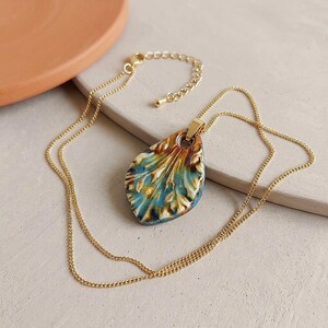 Può includere: Una collana a catena color oro con un ciondolo in ceramica a forma di goccia. Il ciondolo presenta uno smalto lucido con sfumature miste di blu, verde acqua e marrone, mostrando un motivo a foglia testurizzato con rilievi e solchi.