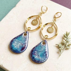Può includere: Un paio di orecchini pendenti color oro con un cerchio e un piccolo ciondolo inciso con un motivo floreale, collegati a un pendente a forma di goccia con un motivo marmorizzato blu, turchese e viola e una trama bianca a ragnatela.