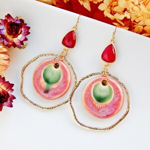 Peut inclure: Une paire de boucles d'oreilles en or avec des cercles en c&eacute;ramique rose et vert et des pierres en forme de larme rouges.