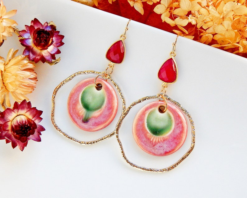 Peut inclure: Une paire de boucles d'oreilles en or avec des cercles en c&eacute;ramique rose et vert et des pierres en forme de larme rouges.