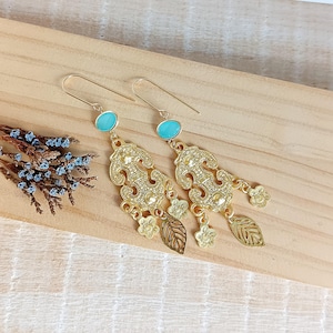 Può includere: Un paio di orecchini pendenti color oro con pietre ovali turchesi e disegni filigranati ornati. Gli orecchini presentano ciondoli a forma di foglie e fiori, esposti su una superficie di legno.