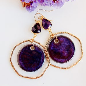 Peut inclure: Une paire de boucles d'oreilles cr&eacute;oles dor&eacute;es avec un pendentif en forme de larme violet et or et un pendentif rond en c&eacute;ramique violet. Le pendentif a une surface textur&eacute;e scintillante.