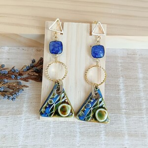 Op de afbeelding: Een paar oorhangers met goudkleurige metalen accenten. Elke oorbel heeft een gouden driehoekige oorbel, een blauw vierkant, een gouden ring en een blauwe, groene en bruine driehoekige hanger. De oorbellen worden gepresenteerd op een houten oppervlak.