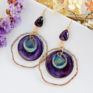 Peut inclure: Boucles d'oreilles violettes et dor&eacute;es avec une pierre pr&eacute;cieuse am&eacute;thyste en forme de larme et un charme rond en c&eacute;ramique avec un motif tourbillonnant.