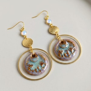 May include: A pair of handcrafted earrings with a celestial theme. Each earring features a gold-colored hoop, a textured gold disc, and a ceramic pendant with a blue and purple glaze. The pendants have a raised floral design and are accented with a small, iridescent bead.
