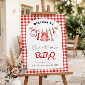 May include: A red and white checkered sign on a wooden easel. The sign reads "Welcome to Olivia + Marcus's BabyQ" with illustrations of baby items. The date "Saturday, July 4, 2026" is printed at the bottom.