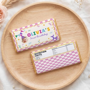 May include: Two rectangular chocolate bars with custom wrappers. One wrapper features a pink and white checkered border, a photo of a baby in a party hat, and the text "OLIVIA'S First Birthday". The other shows a nutrition facts label.