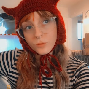May include: A person wearing a red crocheted devil hat with horns and a striped shirt.