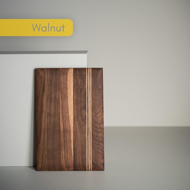 Op de afbeelding: Een rechthoekige walnoten snijplank met een lichte houten accentstreep. De plank heeft een rijke, donkerbruine kleur met lichtere houtnerfpatronen. Het woord "Walnut" wordt weergegeven op een geel label.