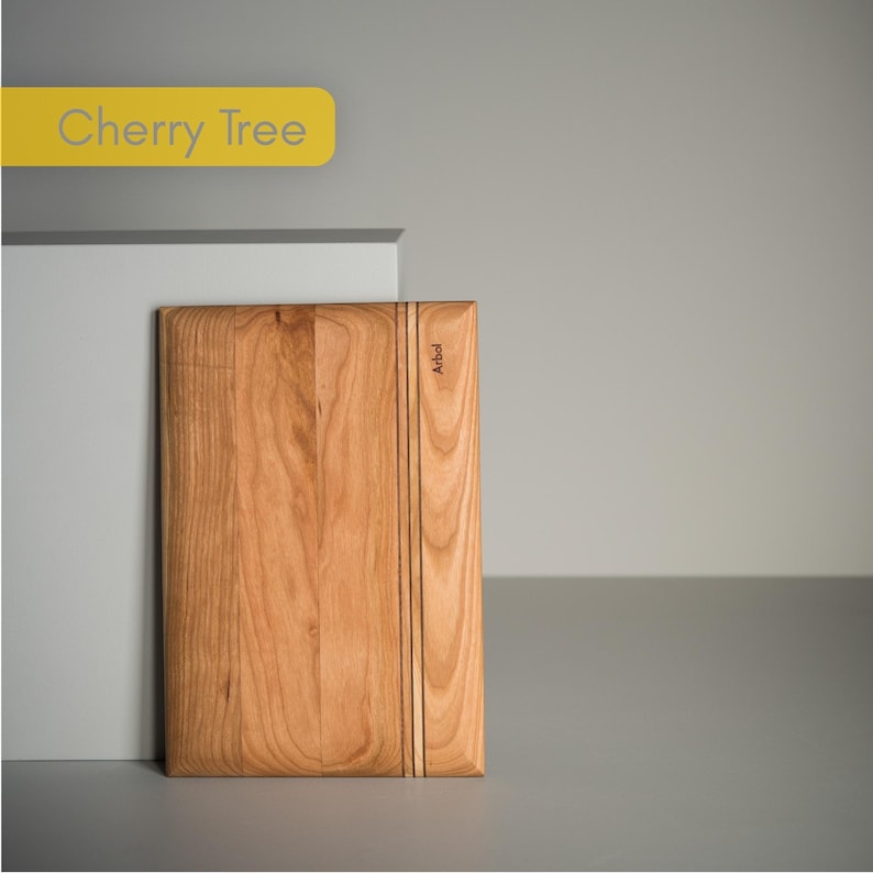 Op de afbeelding: Een rechthoekige snijplank van kersenhout met een natuurlijk houtnerfpatroon. De plank heeft een verticaal designelement met drie donkere lijnen en het woord "Arbol" aan de zijkant. De tekst "Cherry Tree" staat op een gele banner linksboven.