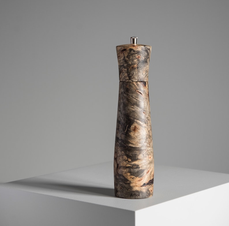 May include: A dark brown wooden pepper mill with a silver top. The mill has a unique wood grain pattern and the word "Arbol" is printed on the bottom.