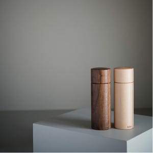 Op de afbeelding: Twee cilindrische houten molens, een gemaakt van donkerbruin notenhout en de andere van lichtbeige esdoornhout. Elke molen heeft een gladde, verticale nerf en een afgeronde bovenkap. Het woord Arbol staat gedrukt op de lichte houten molen.