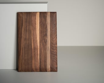 May include: Rectangular wooden cutting board with a dark brown finish. Two thin, light-colored stripes run vertically down the center. The board is made of wood and has a natural grain pattern.