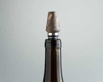 May include: A decorative bottle stopper with a wooden top and a silver-colored metal band. The stopper is on a dark green glass bottle. The wooden top has a textured, brown and gray appearance. The bottle is against a light gray background.
