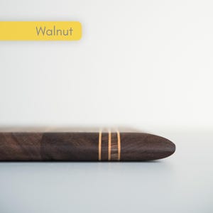 Op de afbeelding: Een donkerbruin houten object met drie lichtgekleurde strepen. Het woord "Walnut" staat in een gele rechthoek linksboven. Het object heeft een gladde, gepolijste afwerking.