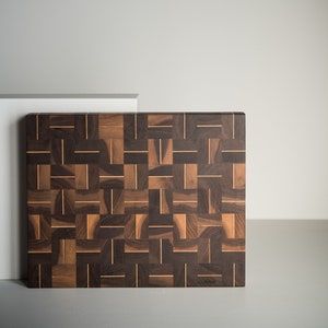 May include: A large, rectangular cutting board made of dark wood with a woven pattern. The board is made of walnut and has a light brown border. The board is signed "Art" in the lower right corner.