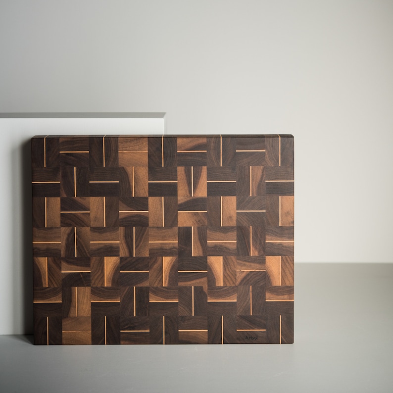 May include: A large, rectangular cutting board made of dark wood with a woven pattern. The board is made of walnut and has a light brown border. The board is signed "Art" in the lower right corner.