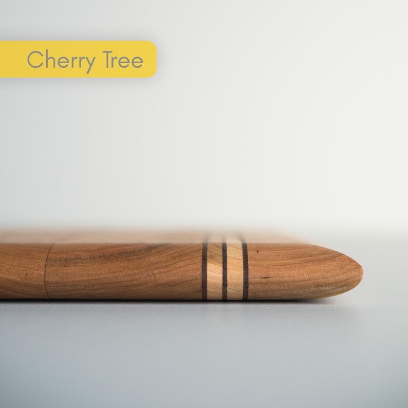 Op de afbeelding: Een houten snijplank van kersenhout met een afgeronde punt. De plank heeft twee dunne, lichtgekleurde strepen met een donkere streep ertussen. De tekst "Cherry Tree" staat in een gele rechthoek linksboven.