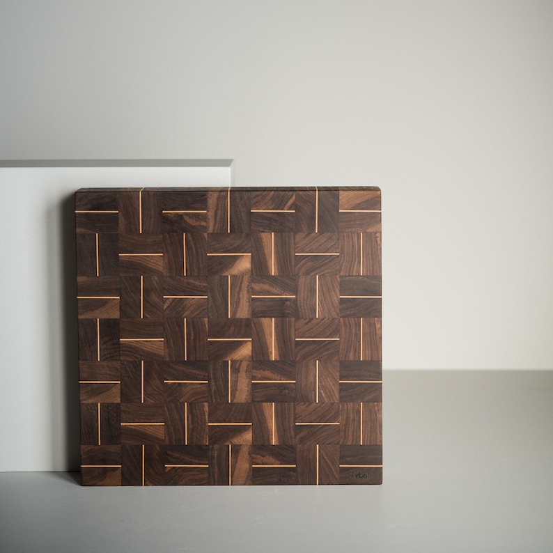 May include: A large, square, wooden cutting board with a woven pattern of dark brown walnut and light brown wood. The board has a natural finish and is made of high-quality wood.