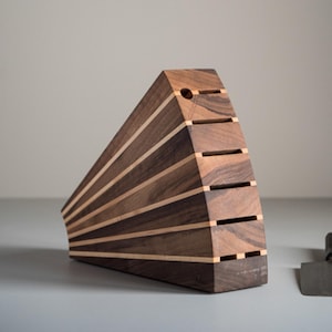 May include: A wooden knife block with a unique, geometric design. The block is made of dark brown and light wood, with horizontal slots for knife storage. A small, metal knife rests beside the block.