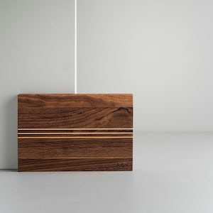 Peut inclure: Une planche à découper rectangulaire en bois de noyer avec deux fines lignes dorées incrustées dans le sens du grain. La planche porte le mot "Arbol" gravé dans le coin inférieur droit.