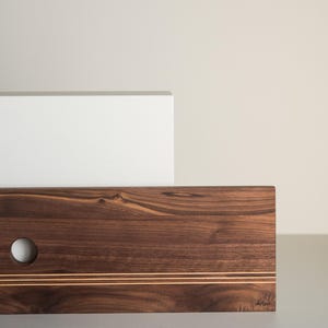 Pode incluir: Uma tábua de corte retangular de madeira com acabamento castanho escuro. Possui um orifício redondo no lado esquerdo e duas incrustações paralelas douradas. A palavra "Arbo" está gravada no canto inferior direito.