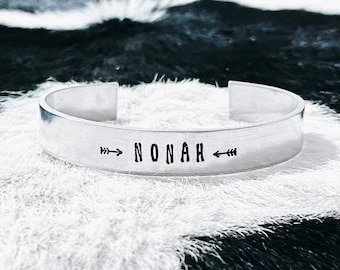 Op de afbeelding: Een zilverkleurige manchetarmband met de naam "NONAH" in zwart gestempeld, geflankeerd door pijldesigns. De open armband ligt op een wit en zwart getextureerd oppervlak.